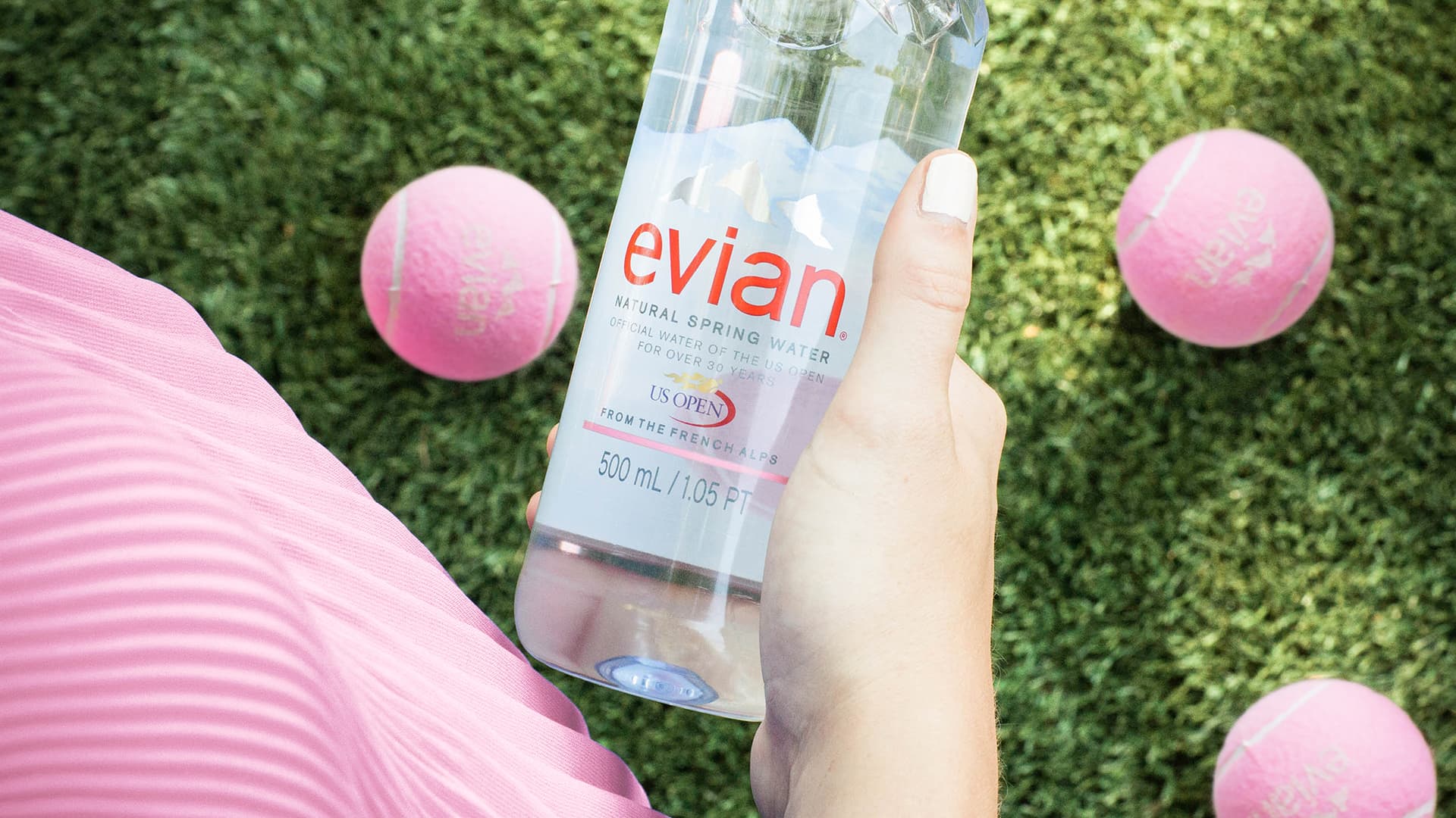 Evian US Open bottle in a woman's hand looking down at pink tennis balls and fresh cut grass